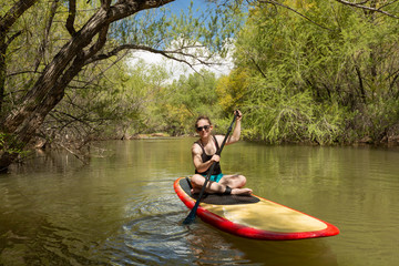 A young woman wearing sunglasses and a swimming suit sits on a paddle board paddling down Quail creek in Southern Utah with willow trees lining the banks on either side. 
