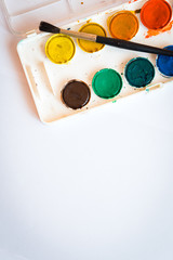 The palette of watercolor paint and one brush on white sheets of paper. Space on left side. Top view. Vertical photo