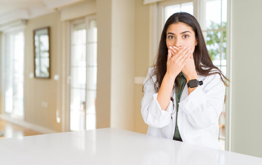 Young woman wearing medical coat at the clinic as therapist or doctor shocked covering mouth with hands for mistake. Secret concept.