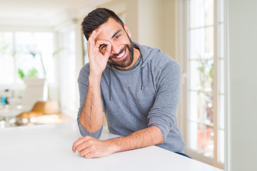 Naklejka na ściany i meble Handsome hispanic man wearing casual sweatshirt at home doing ok gesture with hand smiling, eye looking through fingers with happy face.