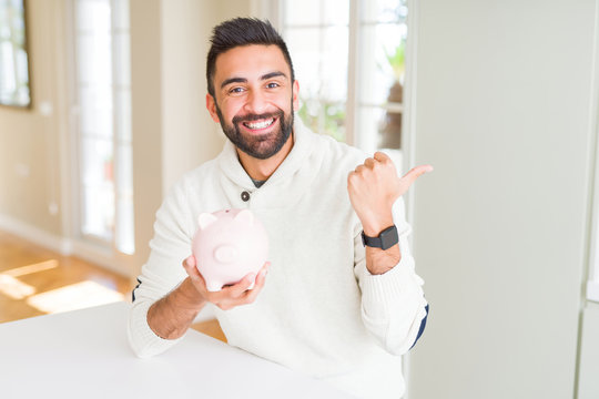 Handsome hispanic man holding piggy bank pointing and showing with thumb up to the side with happy face smiling