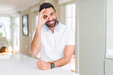 Handsome hispanic man casual white t-shirt at home doing ok gesture with hand smiling, eye looking through fingers with happy face.