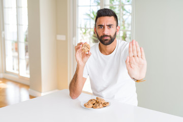 Handsome hispanic man eating chocolate chips cookies with open hand doing stop sign with serious and confident expression, defense gesture