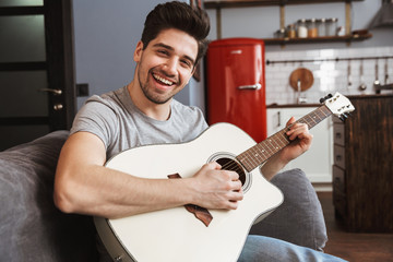 Image of smiling handsome man 30s playing music on acoustic guitar at home