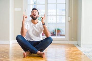 Handsome hispanic man wearing casual t-shirt sitting on the floor at home amazed and surprised looking up and pointing with fingers and raised arms.
