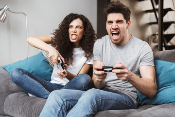 Picture of emotional couple sitting on sofa at home and playing video games together with joysticks