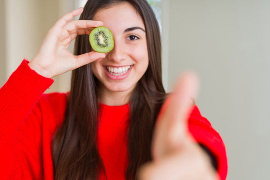 Beautiful young woman eating half fresh green kiwi happy with big smile doing ok sign, thumb up with fingers, excellent sign
