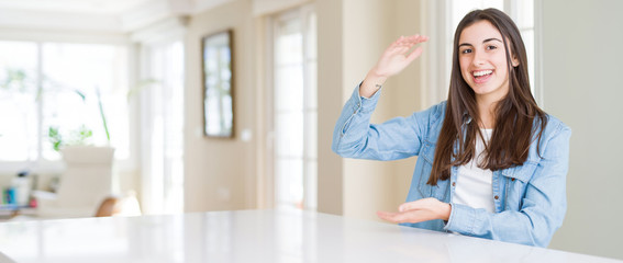 Wide angle picture of beautiful young woman sitting on white table at home gesturing with hands showing big and large size sign, measure symbol. Smiling looking at the camera. Measuring concept.