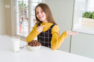 Beautiful young girl kid eating chocolate cereals and glass of milk for breakfast amazed and smiling to the camera while presenting with hand and pointing with finger.