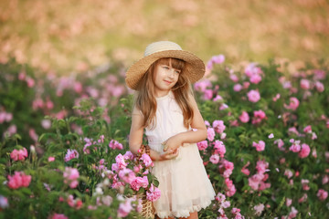 A little girl with beautiful long blond hair, dressed in a light dress and a wreath of real flowers on her head, in the garden of a tea rose
