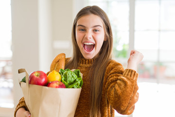 Beautiful young girl holding paper bag of fresh groceries screaming proud and celebrating victory and success very excited, cheering emotion