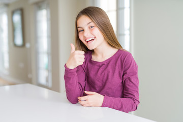 Fototapeta premium Beautiful young girl kid on white table doing happy thumbs up gesture with hand. Approving expression looking at the camera with showing success.
