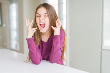 Beautiful young girl kid on white table celebrating mad and crazy for success with arms raised and closed eyes screaming excited. Winner concept