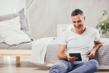 Handsome Caucasian middle-aged man sitting on the floor in bedroom and leaning on bed. In hands tablet.