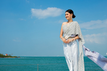 Asian Woman in Blue gray Thai Traditional Costume with jewelry accessories get Winds on Luxury Yacht boat ship in Green Ocean nice sky, Lady relaxing on cruise wearing old formal dress