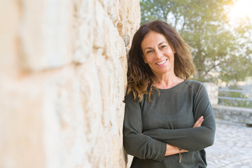 Beautiful middle age woman smiling cheerful leaning on a brick wall at the city street on a sunny day