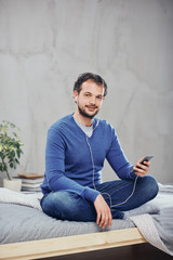 Cute arab man sitting on the bed in bedroom with legs crossed and listening music over smart phone.