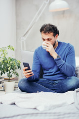 Attractive smiling arab man dressed casual sitting on bed in bedroom, drinking coffee and using smart phone for texting.