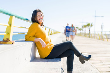 Beautiful young woman sitting on a promenade by the sea smiling cheerful enjoying sunlight relaxing on sunny day