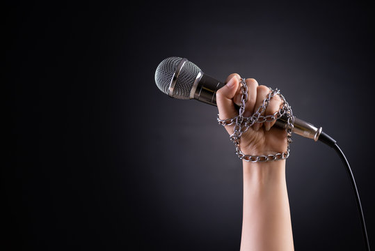 Woman hand with microphone tied with a chain, depicting the idea of freedom of the press or freedom of expression on dark background. World press freedom day concept.