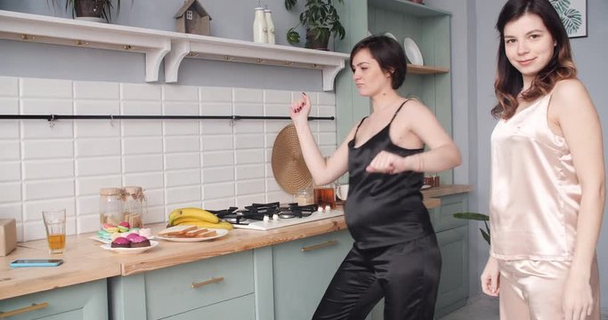 Two attractive young pregnant girls in silk pyjamas having fun, dancing and eating delicious cupcakes together in stylish modern kitchen. Babyshower.