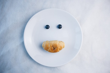 fresh blueberry berries and croissant in the form of a face with a smileon a white plate close-up. breakfast of wild berries. copy space