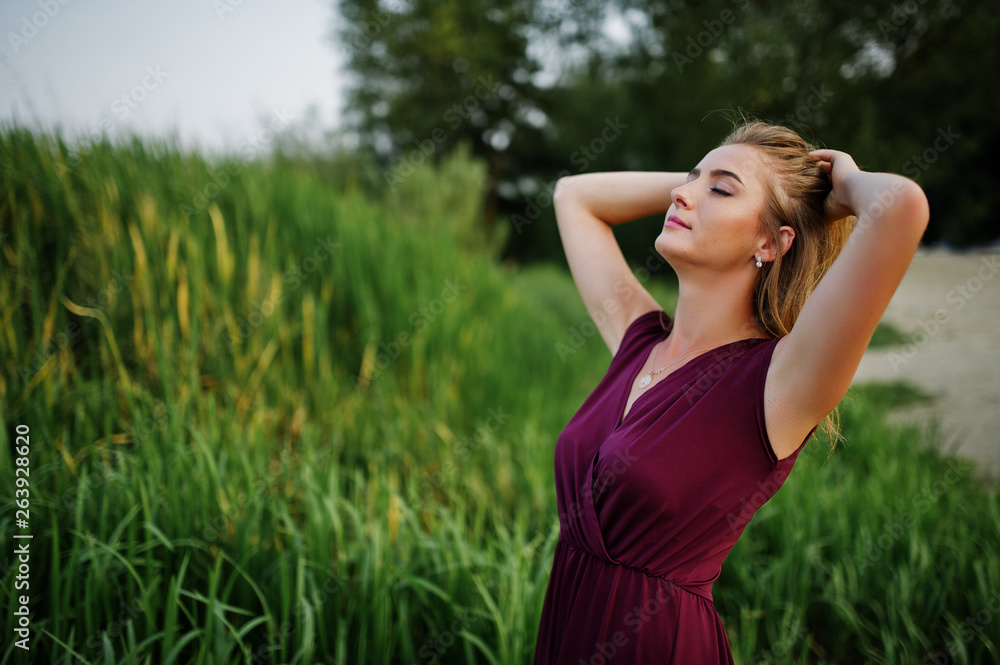 Wall mural Blonde sensual woman in red marsala dress posing against lake with reeds.