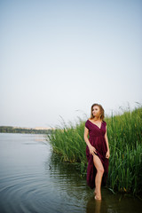 Fototapeta premium Blonde sensual woman in red marsala dress standing in water of lake with reeds.