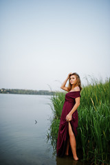 Obraz premium Blonde sensual woman in red marsala dress standing in water of lake with reeds.