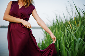 Blonde sensual woman in red marsala dress standing in water of lake with reeds.