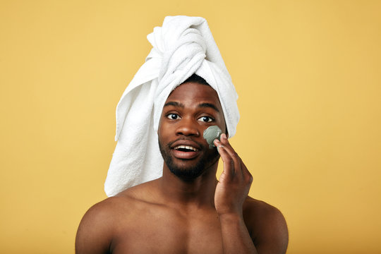 Young happy man with a towel on his head applying mask on his face, close up photo, daily routine concept. health care