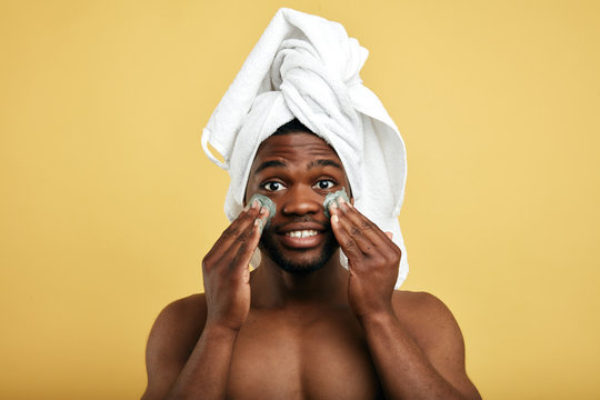 puzzled man with palms on his cheek and green cream posing to the camera, isolated yellow background. man is allergic to the cosmetic, skin problems