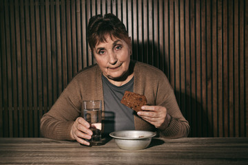 Poor senior woman with water and bread at table against wooden wall
