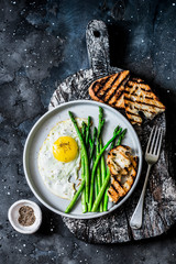 Simple rustic breakfast - fried egg, asparagus and grilled bread on a dark background, top view