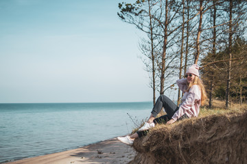 Young adult woman in pink hat sitting alone on the bluffs, looking of sea, freedom concept, peaceful atmosphere, meditation, copy space, Latvia, Jurkalne