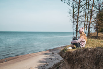 Young adult woman in pink hat sitting alone on the bluffs, looking of sea, freedom concept, peaceful atmosphere, meditation, copy space, Latvia, Jurkalne