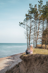Obraz premium Young adult woman in pink hat sitting alone on the bluffs, looking of sea, freedom concept, peaceful atmosphere, meditation, copy space, Latvia, Jurkalne