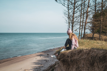 Young adult woman in pink hat sitting alone on the bluffs, looking of sea, freedom concept,...