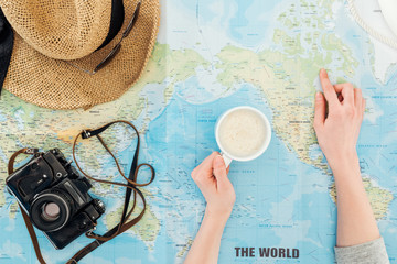Cropped view of woman with cup of cappuccino, film camera, sunglasses and straw hat on world map