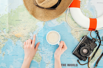Fototapeta na wymiar Cropped view of woman with cup of cappuccino, film camera, sunglasses and straw hat pointing with finger on world map