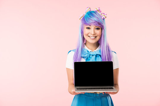 Smiling asian anime girl in wig showing laptop with blank screen isolated on pink
