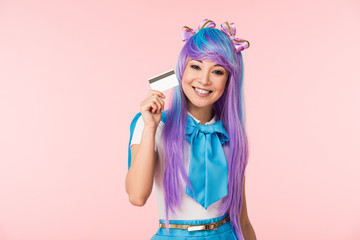 Smiling asian anime girl in purple wig holding credit card isolated on pink