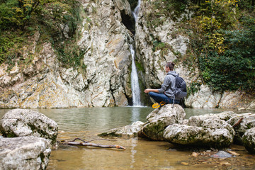 Young male tourist in casual clothes with backpack sits in picturesque place on stone overlooking...