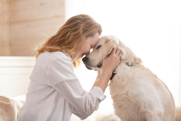 Young pretty woman in casual clothes hugging her beloved big white dog sitting on the sofa in the living room of her cozy country house. Animal communication concept