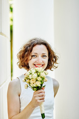 Portrait of smiling young woman with red lipstick in white dress outdoor. Curly brown hair bride with a wedding bouquet