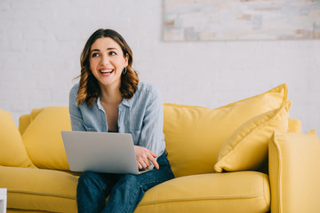 Smiling pretty freelancer sitting on yellow sofa with laptop