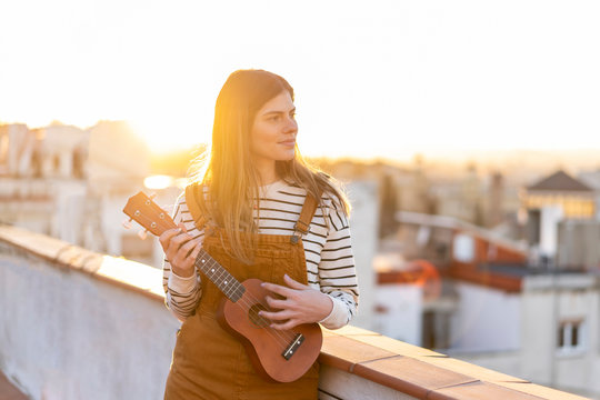 Young woman standing on roof terrace in the evening playing ukulele