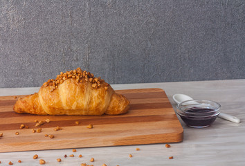 One croissant on a wooden cutting board, sprinkled with nuts on a light wooden table. Nuts scattered on the table surface. Cup with blueberry jam and a teaspoon. On a gray background.