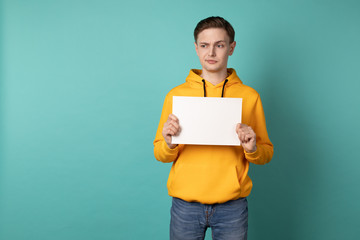 Confused unhappy young man in yellow hoodie standing against blue wall