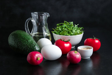 ingredients for healthy salad on black background.boiled eggs avocado radish arugula tomatoes olive oil sesame seeds